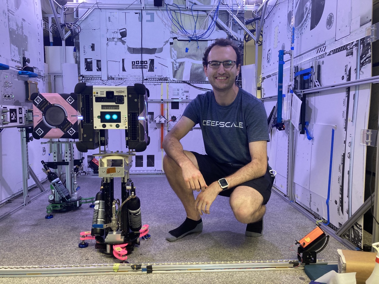 Picture of me kneeling near one of the cube-shaped astrobee robots in the testing lab. The robot is mouted on a floatation platform.